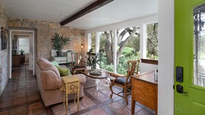 A cozy living room with a stone wall, a wooden ceiling, a green door, and a view of trees through large windows.