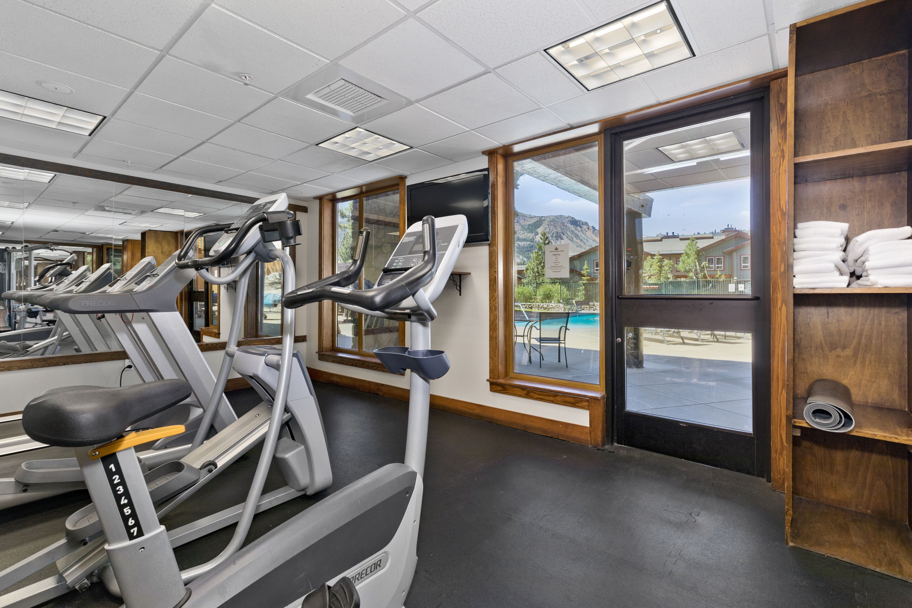 Un gimnasio con cintas de correr, vistas a la piscina y un estante de madera con toallas.