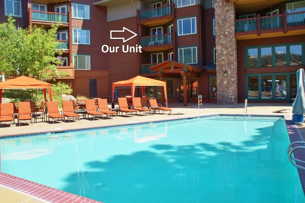 Un espace piscine d’hôtel avec des chaises longues et des parasols, un pilier en pierre et un immeuble à plusieurs étages doté de balcons.