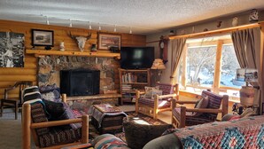 Interior acogedor de una cabaña de troncos con chimenea de piedra, muebles de madera y un amplio ventanal con vistas a un paisaje nevado.