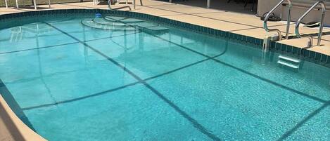 A clear swimming pool with a covered patio area, lounge chairs, and a view of the building in the background.