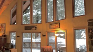 Una sala de estar acogedora con chimenea, ventanas amplias con vista a una terraza nevada y un cartel que indica vistas al río Truckee.
