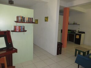 A room with a bookshelf, a desk, a chair, a small table, and a kitchen area in the background.