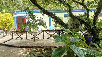 A tropical garden with a wooden deck, a red door, and a blue building in the background.