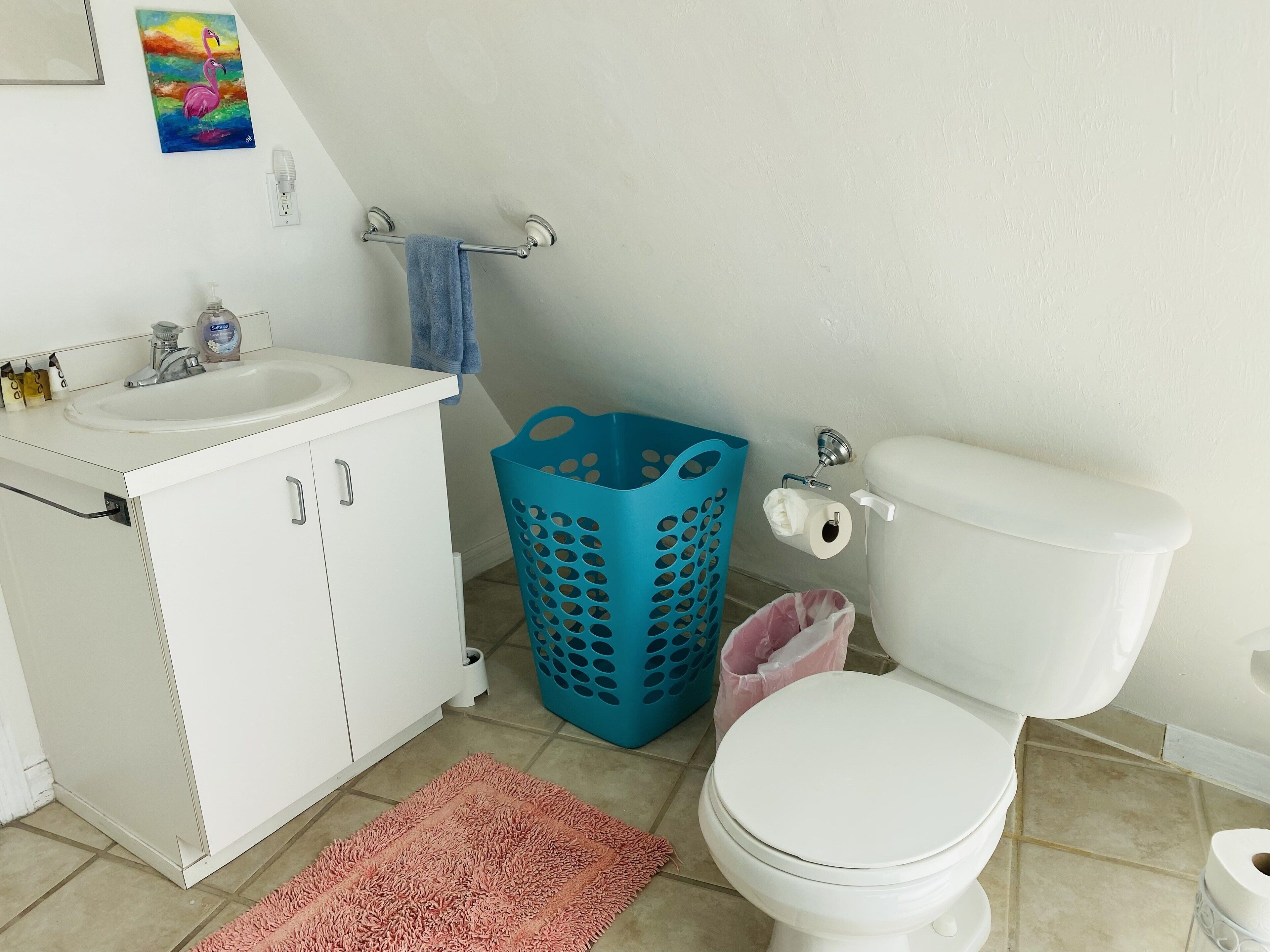 Ein Badezimmer mit Waschbecken, Schrank und Toilette in Weiß. Auf dem Boden steht ein blauer Wäschekorb und ein rosafarbener Müllbeutel.