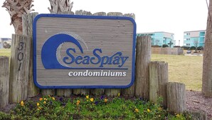 En träskylt med texten ”SeaSpray – bostadsrätter” omgiven av ett trästaket och blommor.