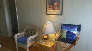 Ett rum med en flätad fåtölj, ett litet bord med en lampa och en inramad tavla på väggen.