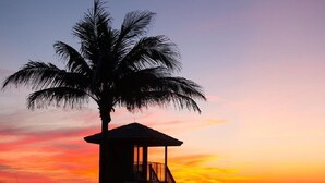 Un cabanon de plage avec un escalier, un palmier et un coucher de soleil.