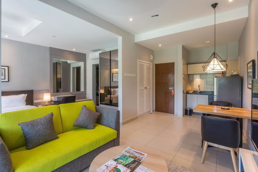 Une chambre d’hôtel moderne avec un canapé vert vif, un coin repas doté d’une table et de chaises en bois, un coin cuisine avec un luminaire suspendu et un grand miroir qui reflète la pièce.