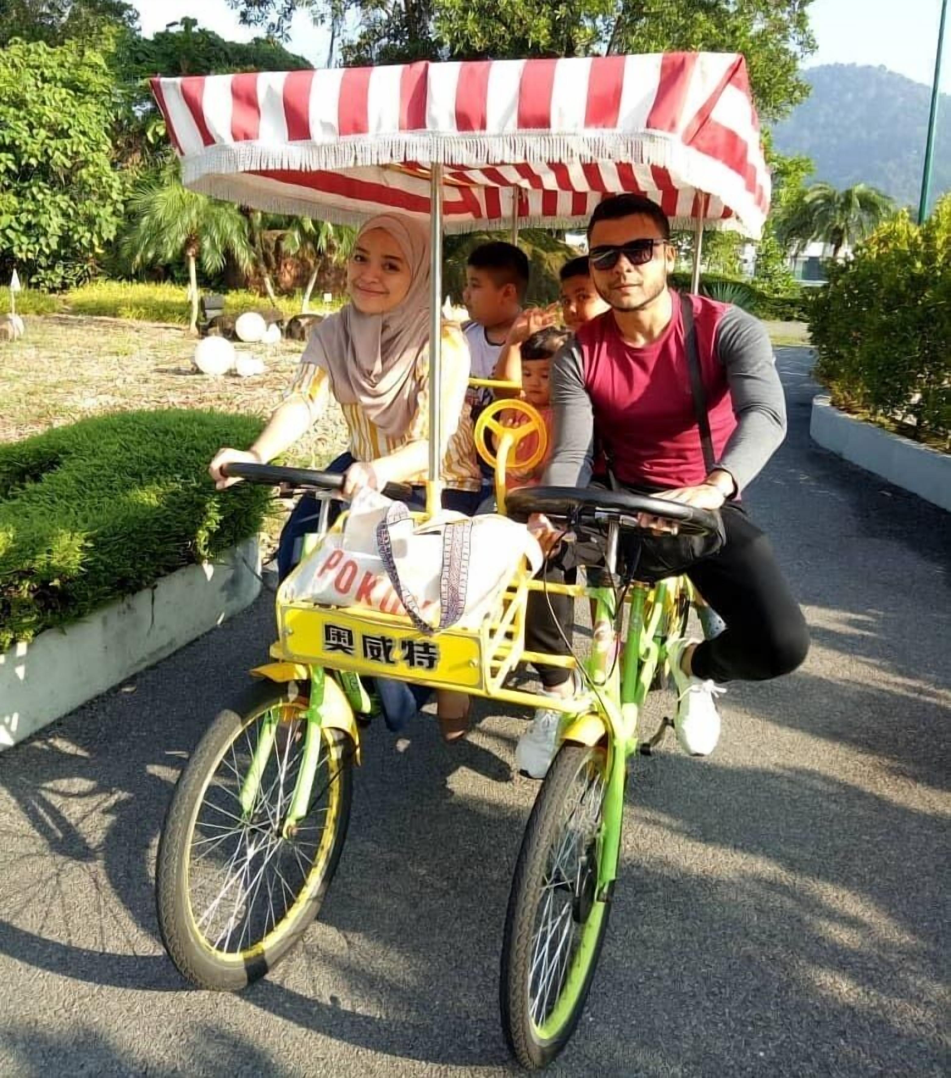 Ein gelber Fahrradwagen mit einem rot-weiß gestreiften Verdeck, beschriftet mit 'POK' und chinesischen Schriftzeichen, wird von zwei Erwachsenen und einem Kind gefahren.