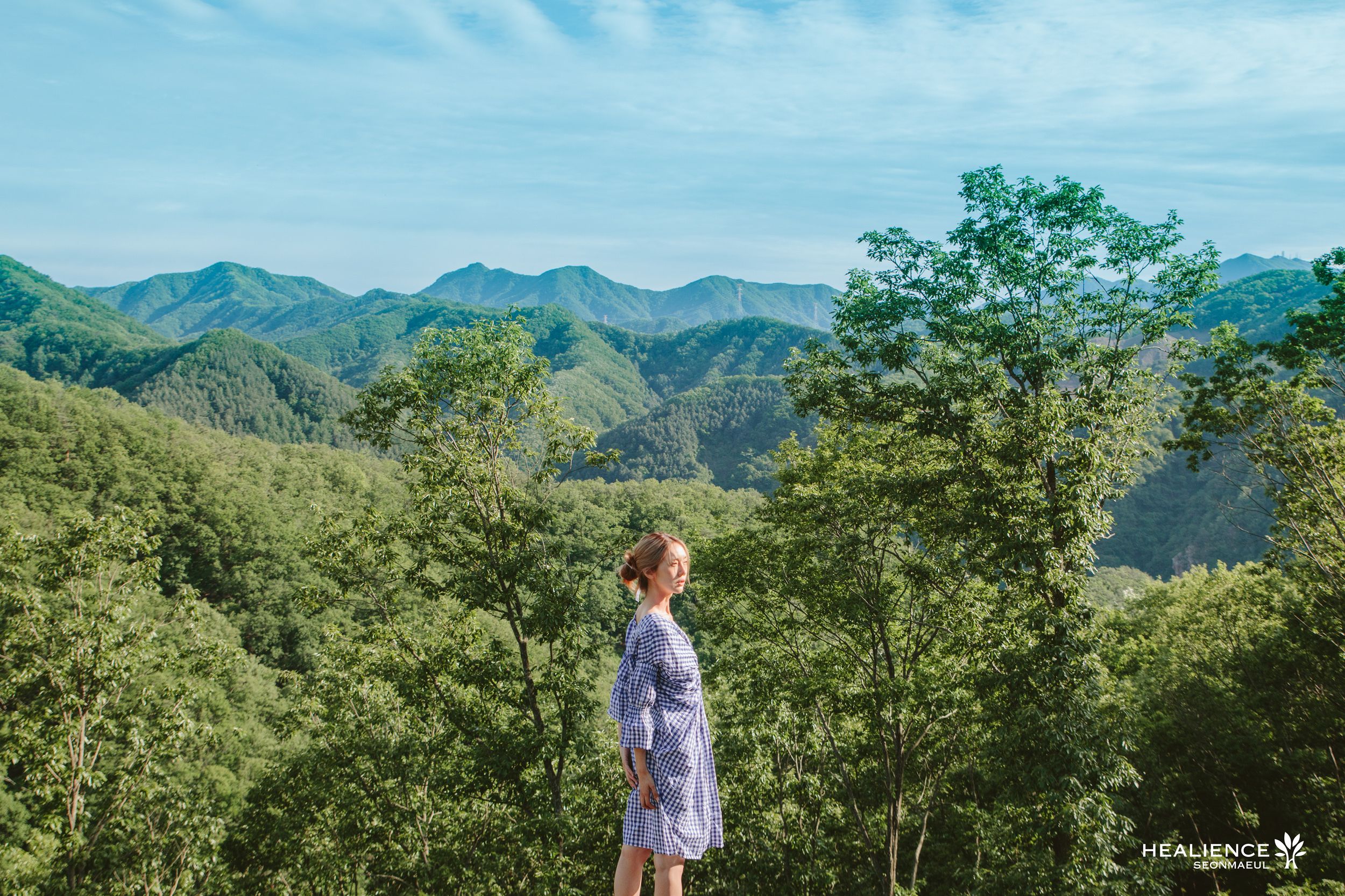A person standing in a forested area with mountains in the background.