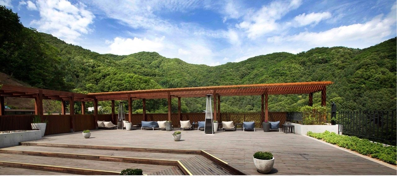 A wooden deck with outdoor seating and a pergola, surrounded by lush greenery and hills.