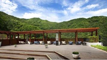 A wooden deck with outdoor seating and a pergola, surrounded by lush greenery and hills.