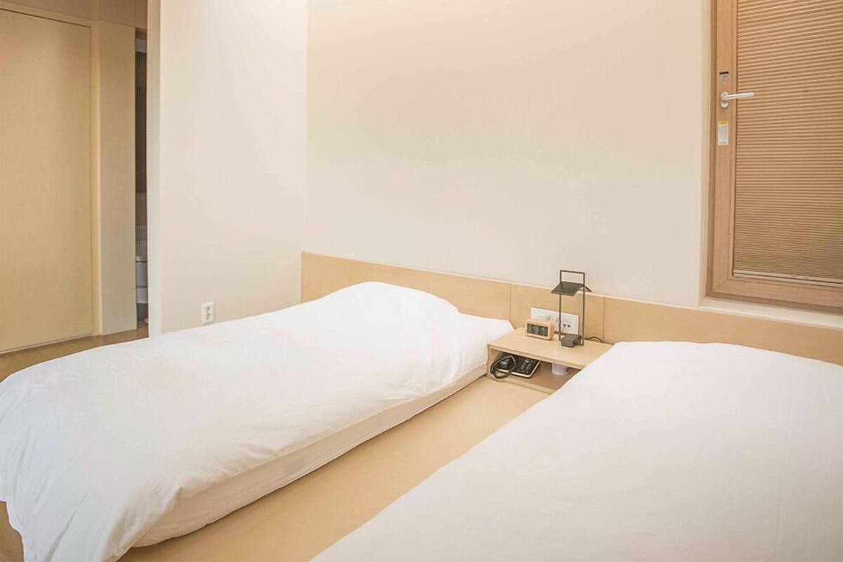 A hotel room with two single beds, a wooden headboard, a bedside table with a lamp, and a window with a view.