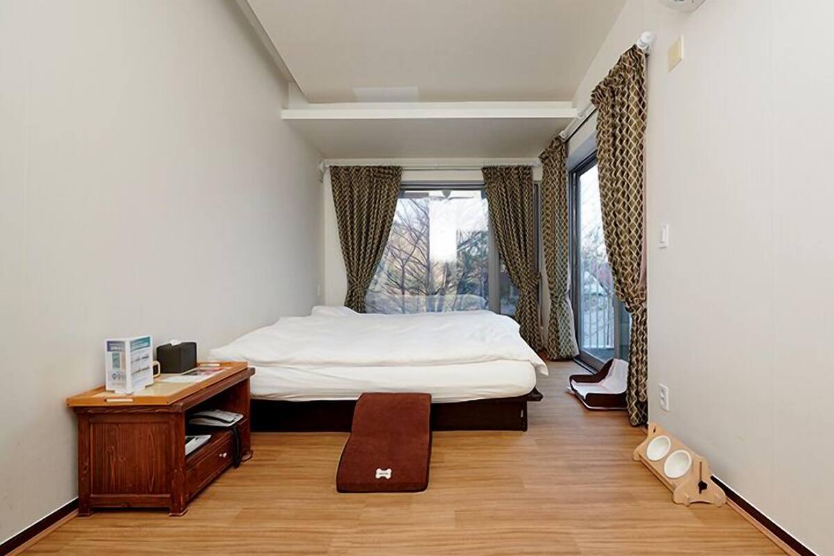 A small, modern bedroom with a bed, a wooden nightstand, a pet bed, and a pet feeding station.
