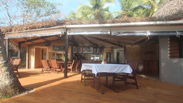 Una terraza exterior de madera con mesas y sillas, un espacio de estar cubierto y una barra con menú.
