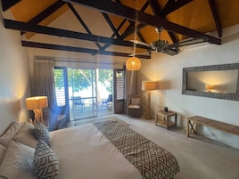 A bedroom with a large bed, a desk, a chair, a small table, and a ceiling fan. There is a large window with curtains and a view of the outdoors.