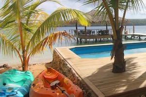 A wooden deck with a pool, palm trees, and kayaks.