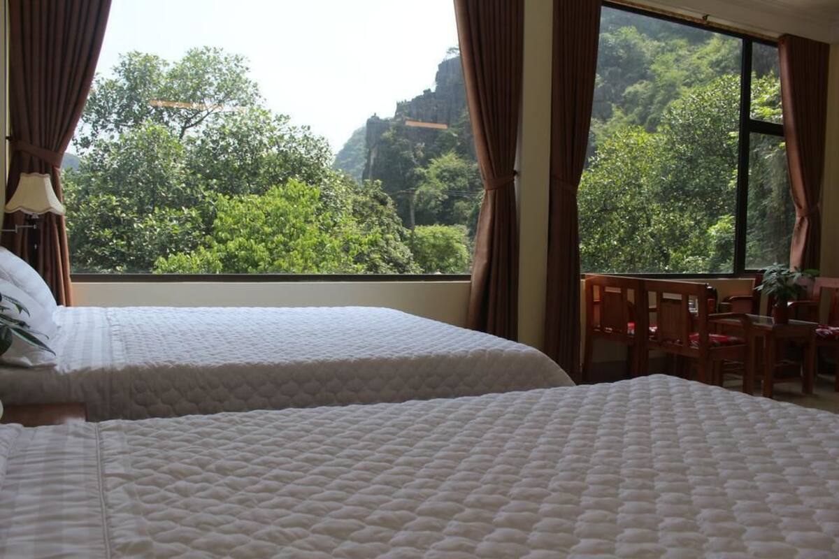 A hotel room with two beds, a dining table, and a view of lush greenery and a mountain.