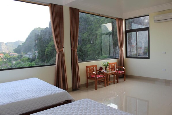 Habitación de hotel con dos camas, una mesa de comedor con sillas y ventanas amplias con vistas a un paisaje montañoso.