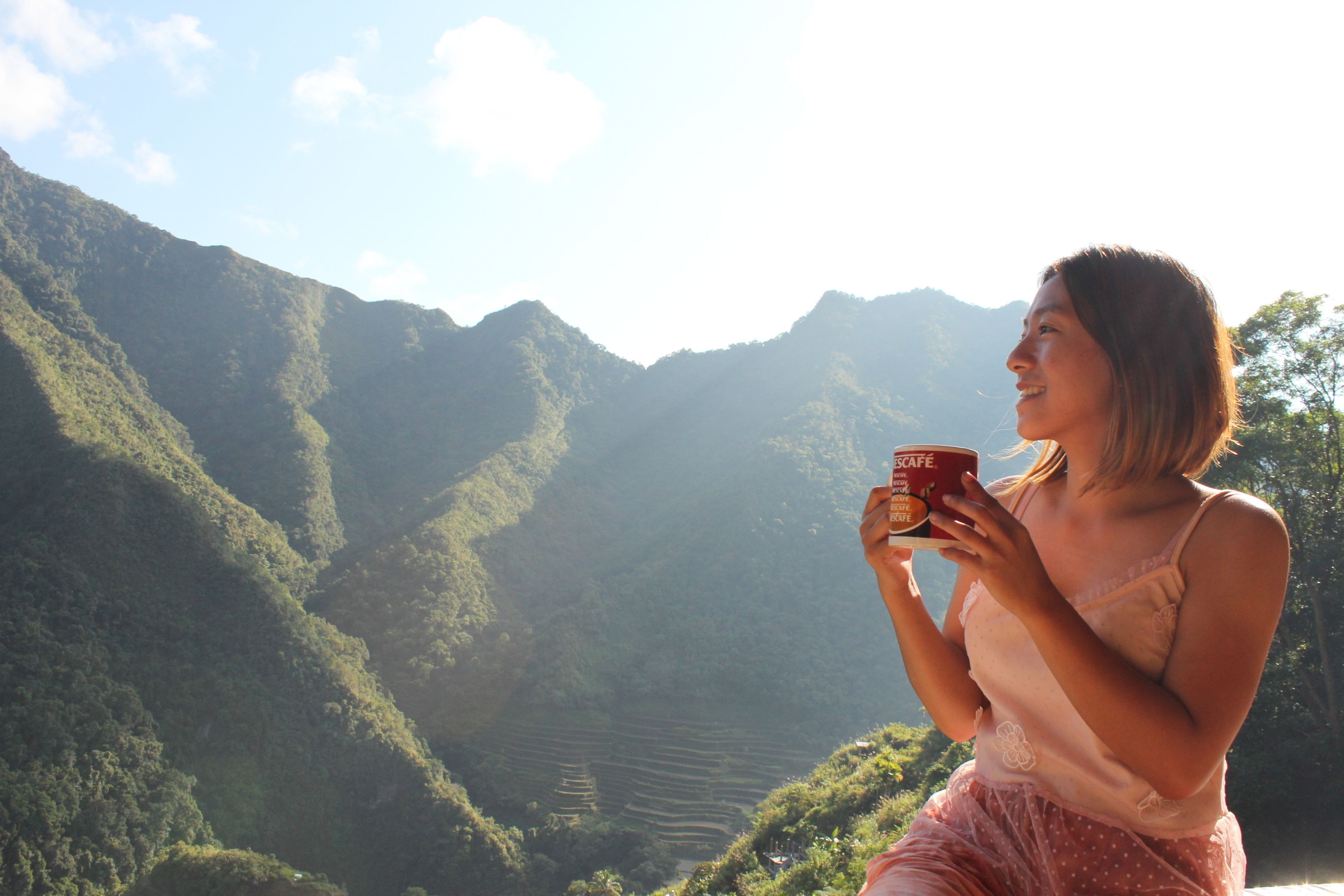 Una persona sosteniendo una taza, observando un paisaje montañoso con campos en terrazas.