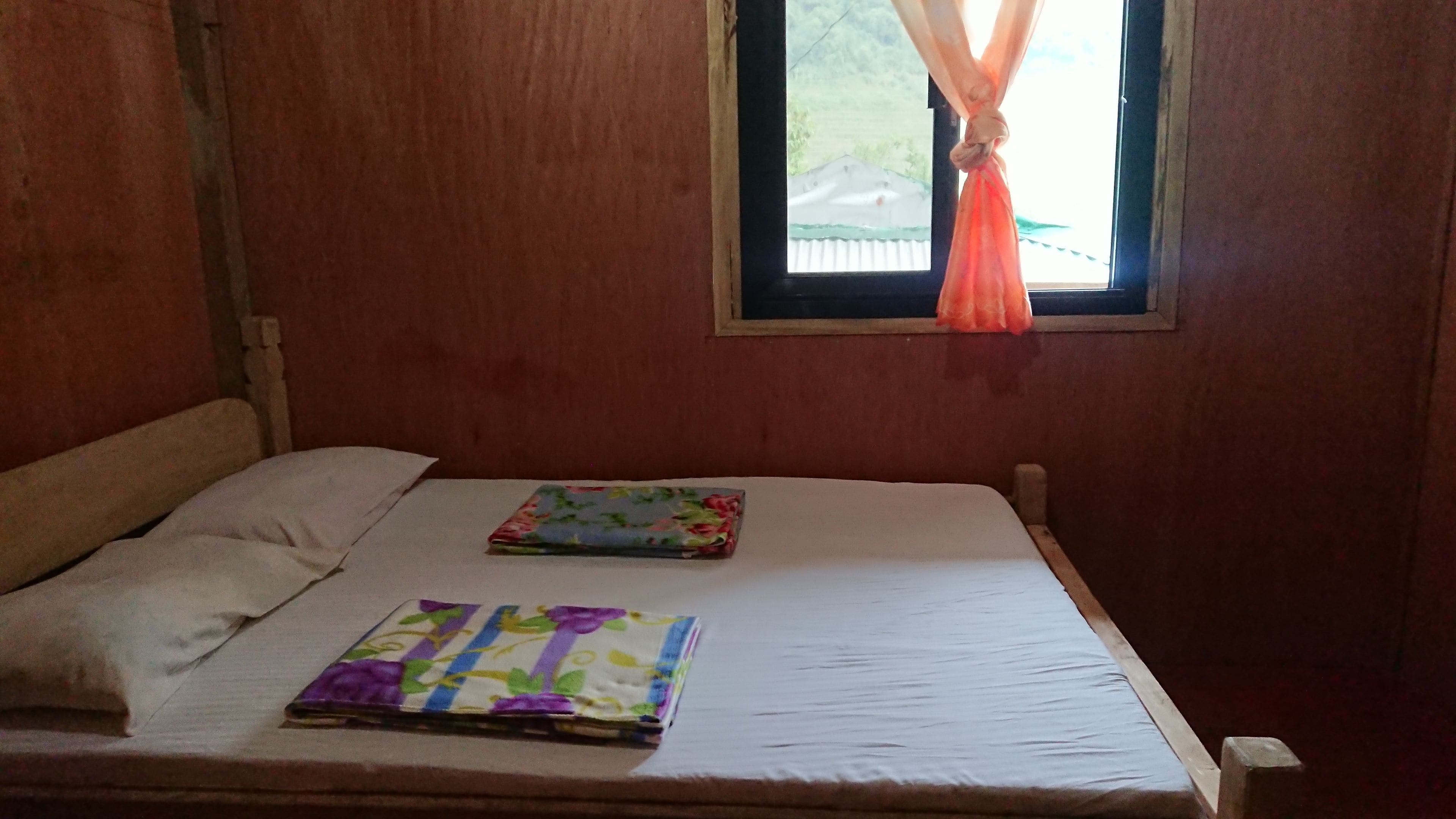 A single bed with a plain mattress and a pillow, a window with a curtain, and a small table with a colorful cloth.