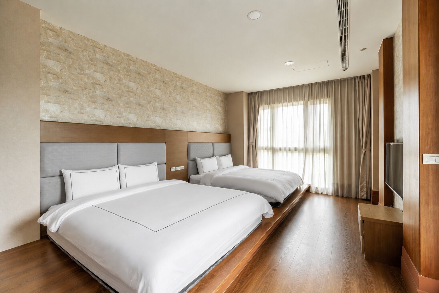 A hotel room with two beds, a large window with curtains, a flat-screen TV, and a wooden floor.