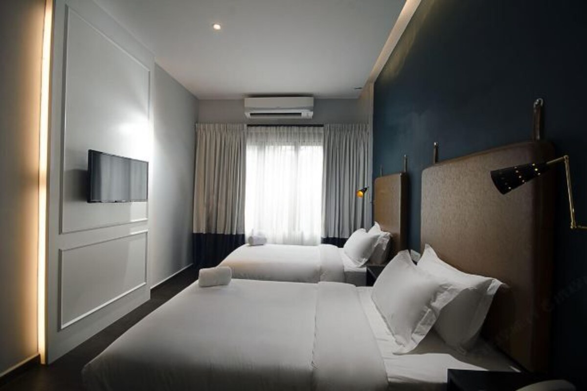 A hotel room with two beds, a flat-screen TV mounted on the wall, a window with curtains, and an air conditioning unit.