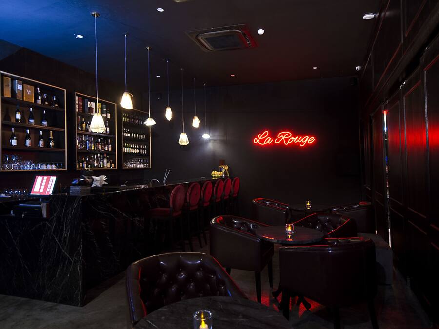 Zona de bar con un letrero de neón que dice "La Rouge", estantes llenos de botellas y asientos de cuero.