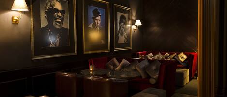 A dimly lit lounge area with framed portraits on the wall, red upholstered seating, and small tables.