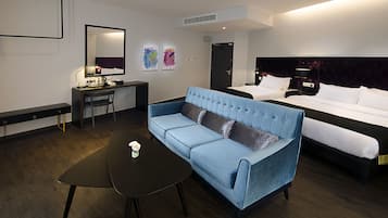 A hotel room with a blue sofa, a small table with a vase, a bed with white linens, and a desk with a chair.