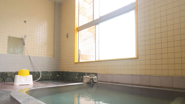 Un petit bain à remous rectangulaire, avec un seau jaune et un tuyau, placé devant un mur carrelé orné d’une fenêtre.
