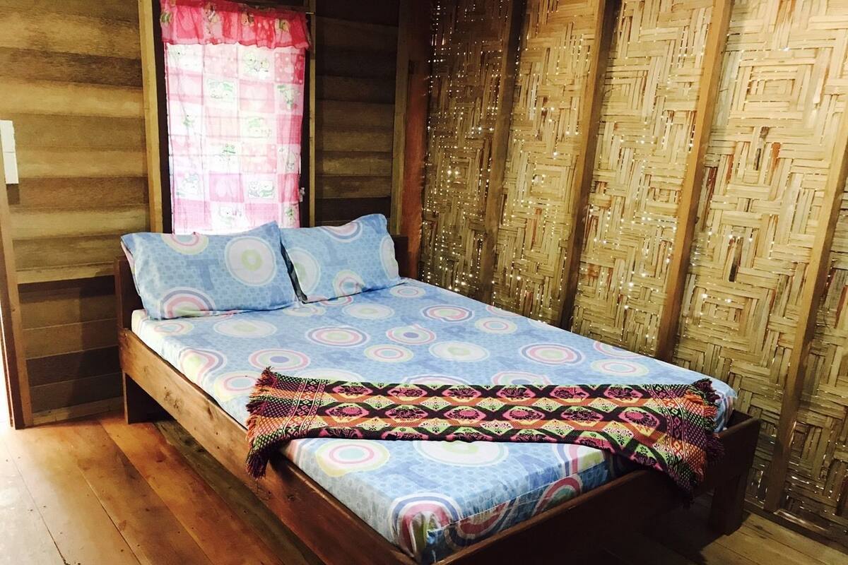 A wooden cabin-style room with a bed, a window with a pink curtain, and a woven wall panel.