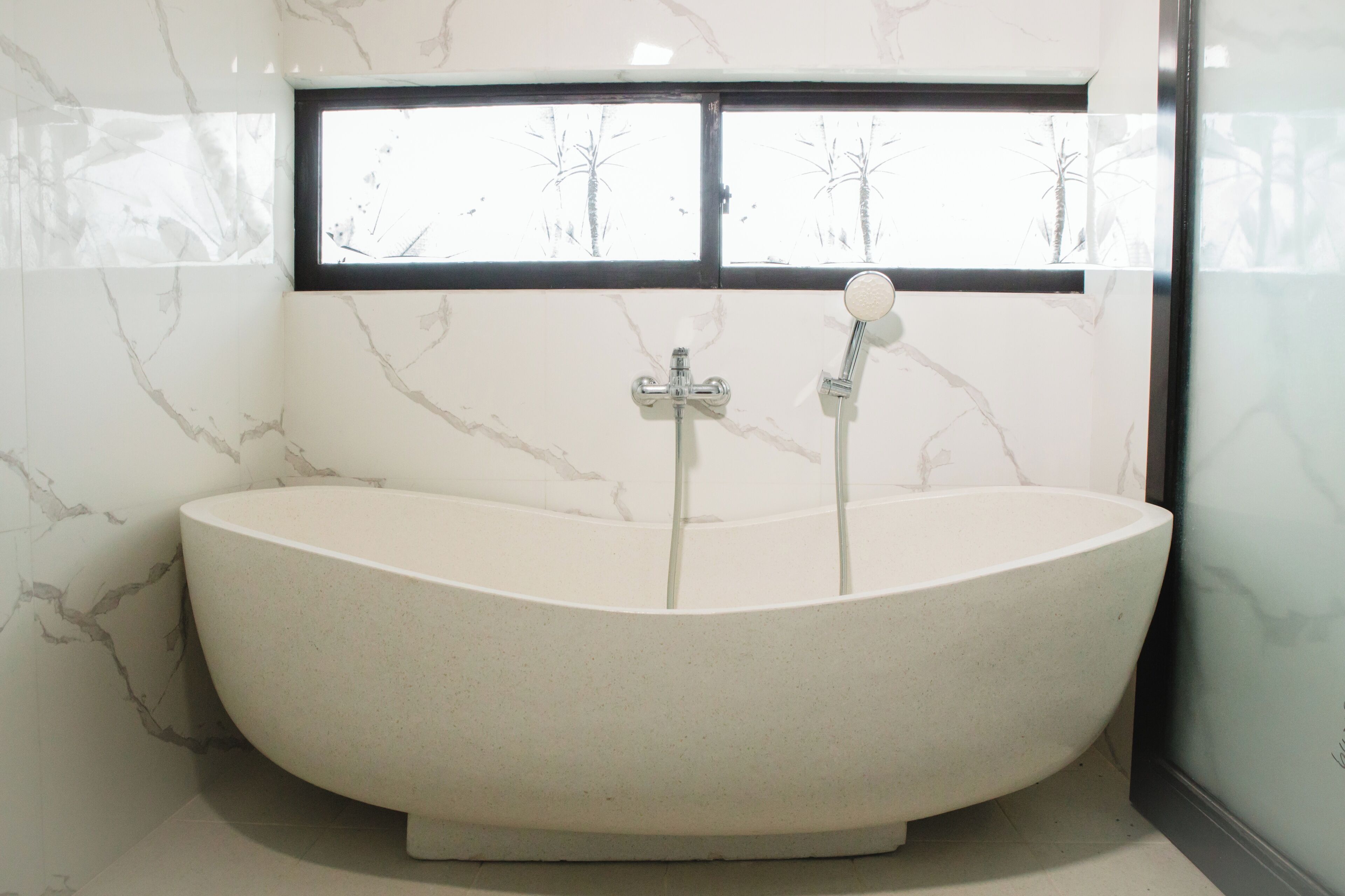 A freestanding bathtub in a bathroom with marble walls and a window above it.