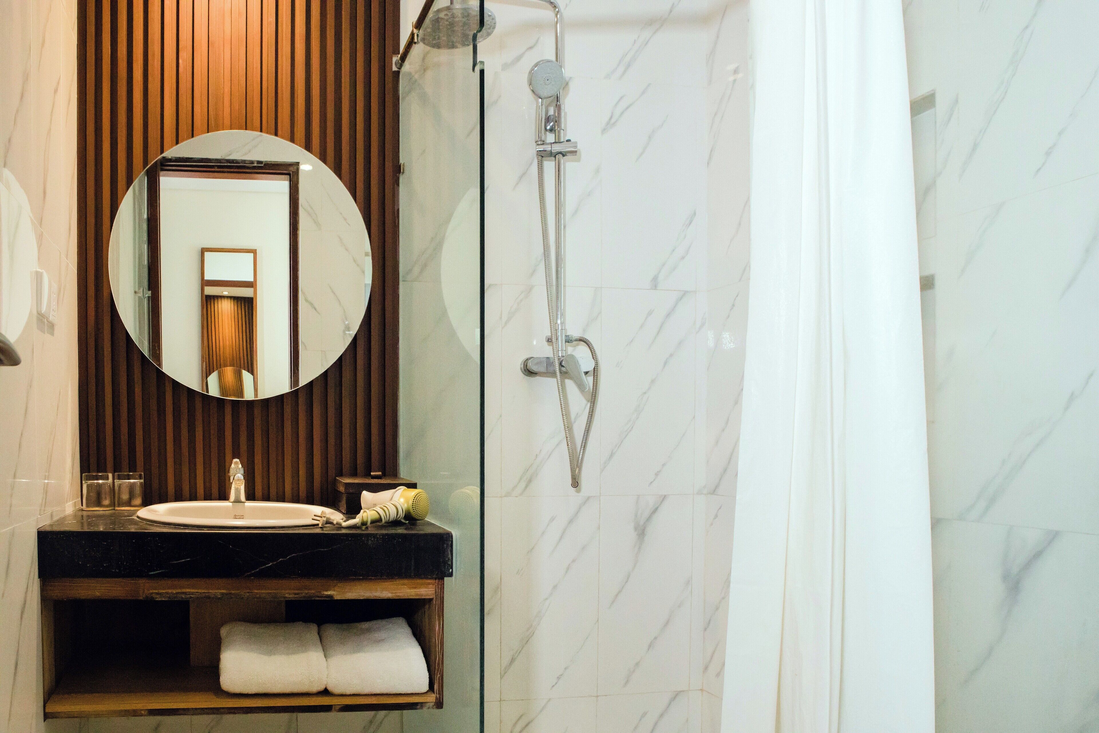 A bathroom with a round mirror, a sink, a shower, and a towel rack.