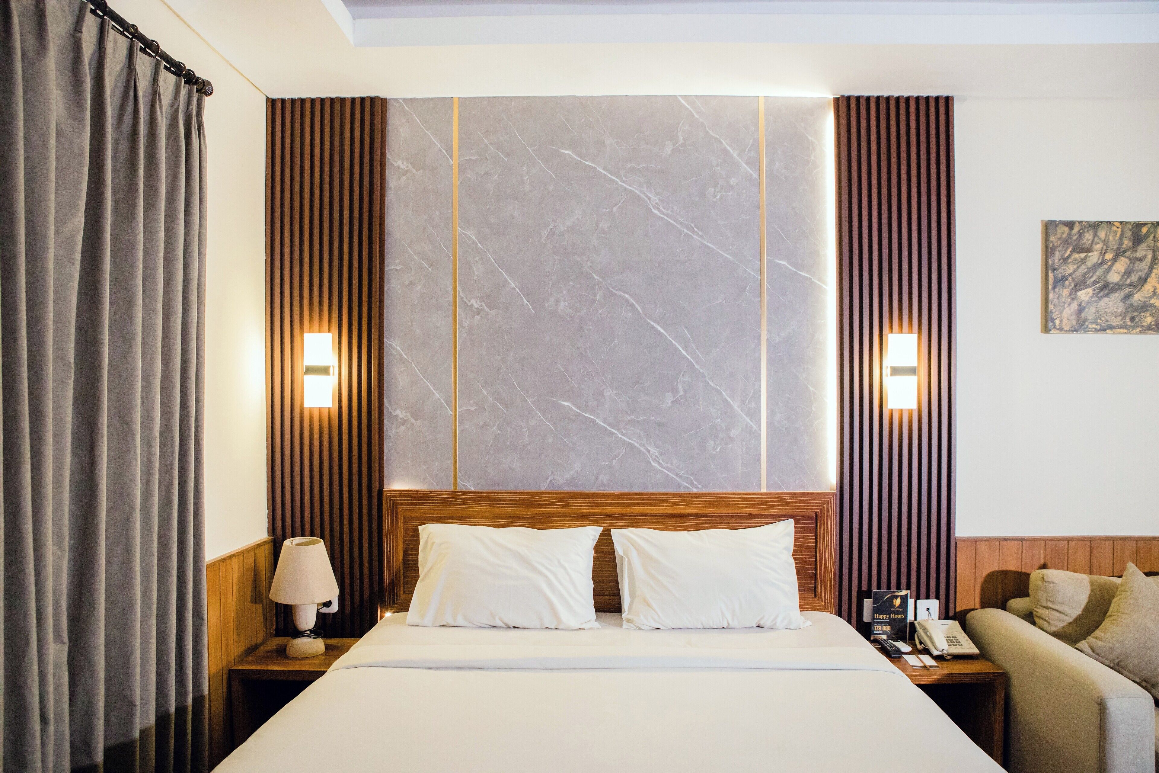 Quarto de hotel com cama, mesa de cabeceira com abajur, sofá, parede com persianas verticais e parede de mármore.