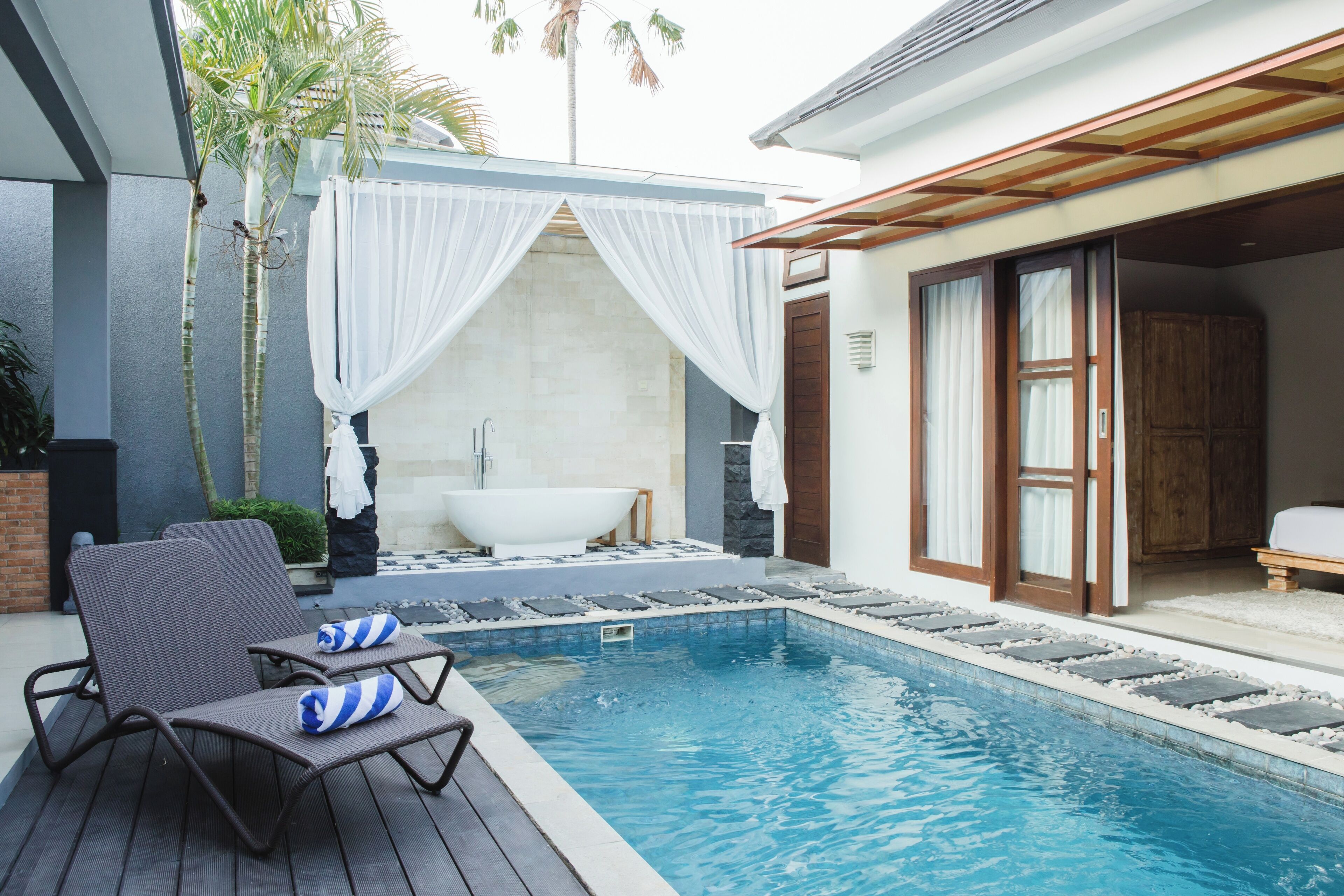 Piscina con sillas de descanso y bañera, rodeada por una terraza de madera y un dosel blanco.