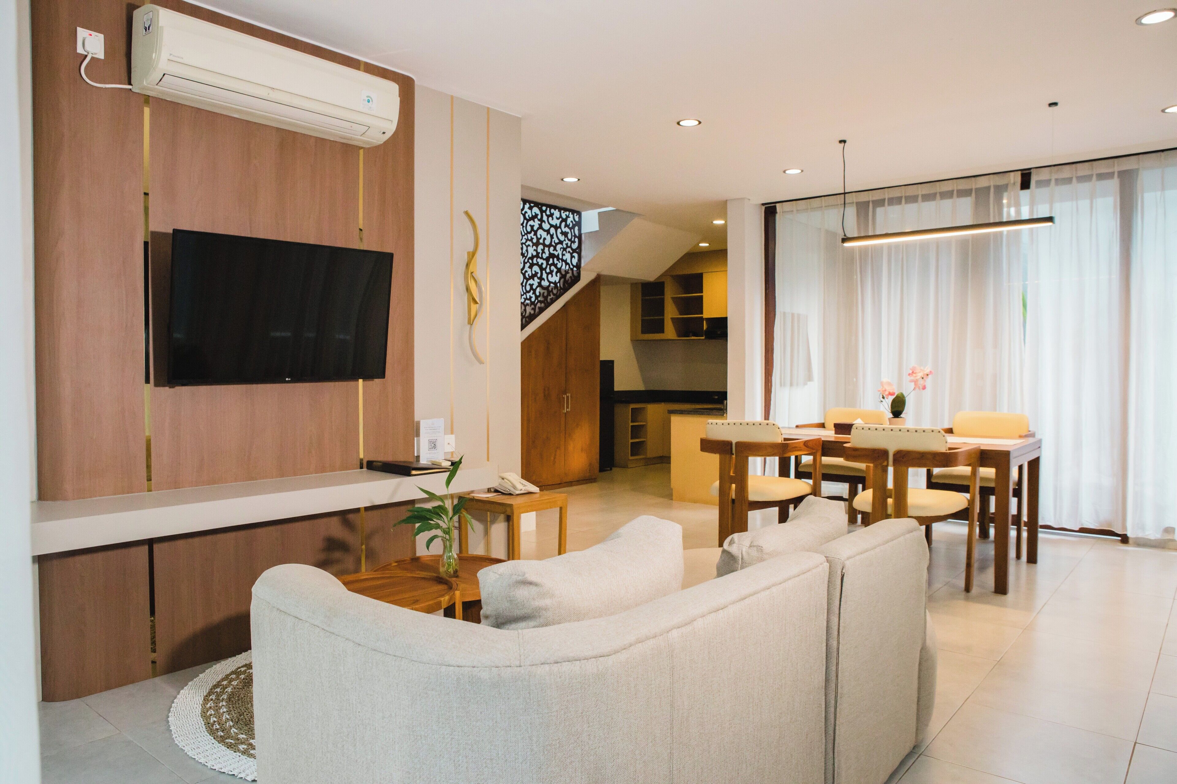 Uma sala de estar moderna com televisão de tela plana, um sofá, uma mesa de jantar com cadeiras e uma cozinha ao fundo.