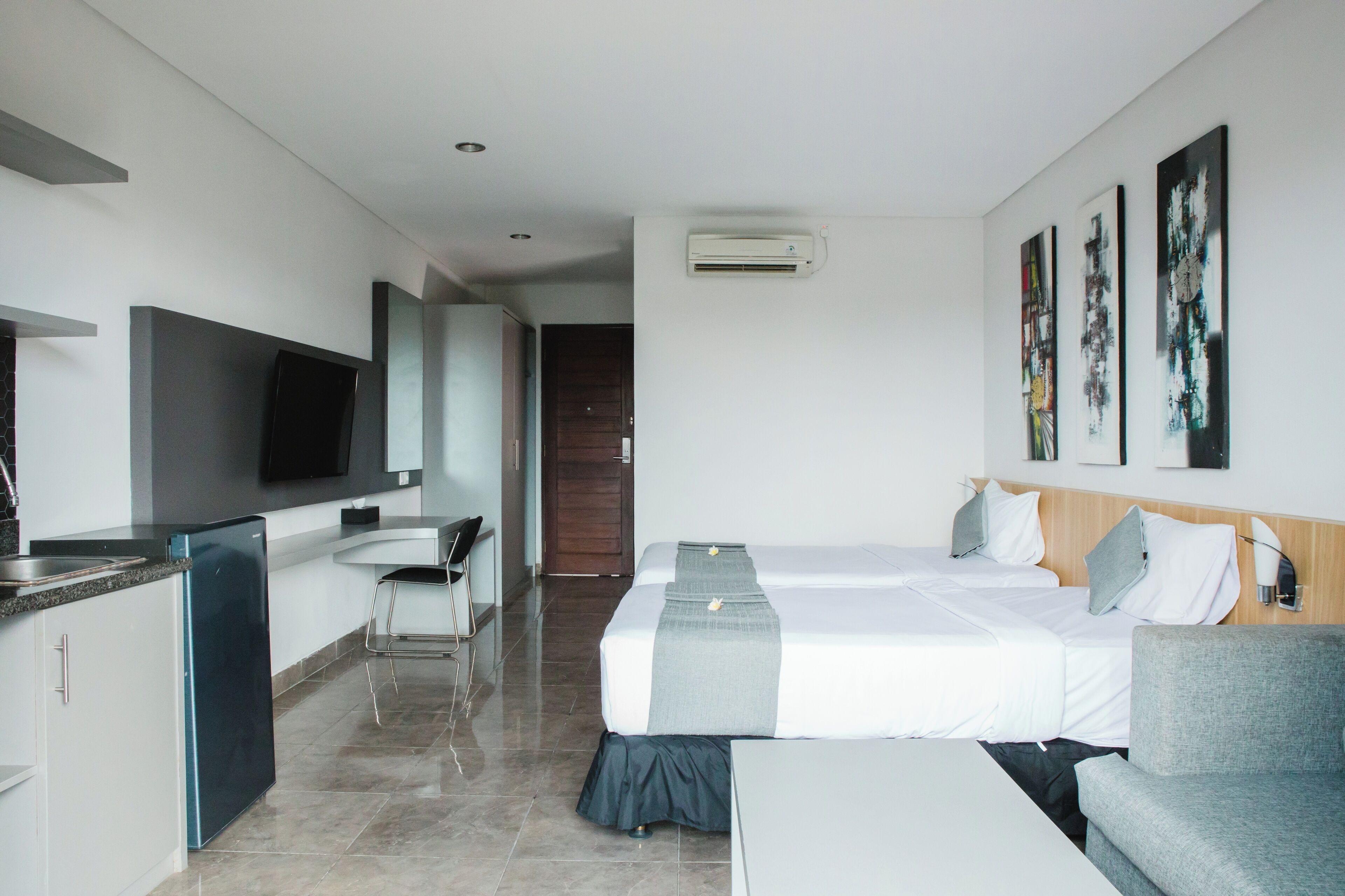 A hotel room with a bed, a desk, a chair, a small kitchenette, and artwork on the walls.
