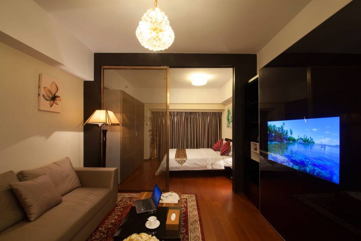 A hotel room with a sofa, a desk with a laptop, a television displaying a tropical scene, and a bed with a red and white pillow.