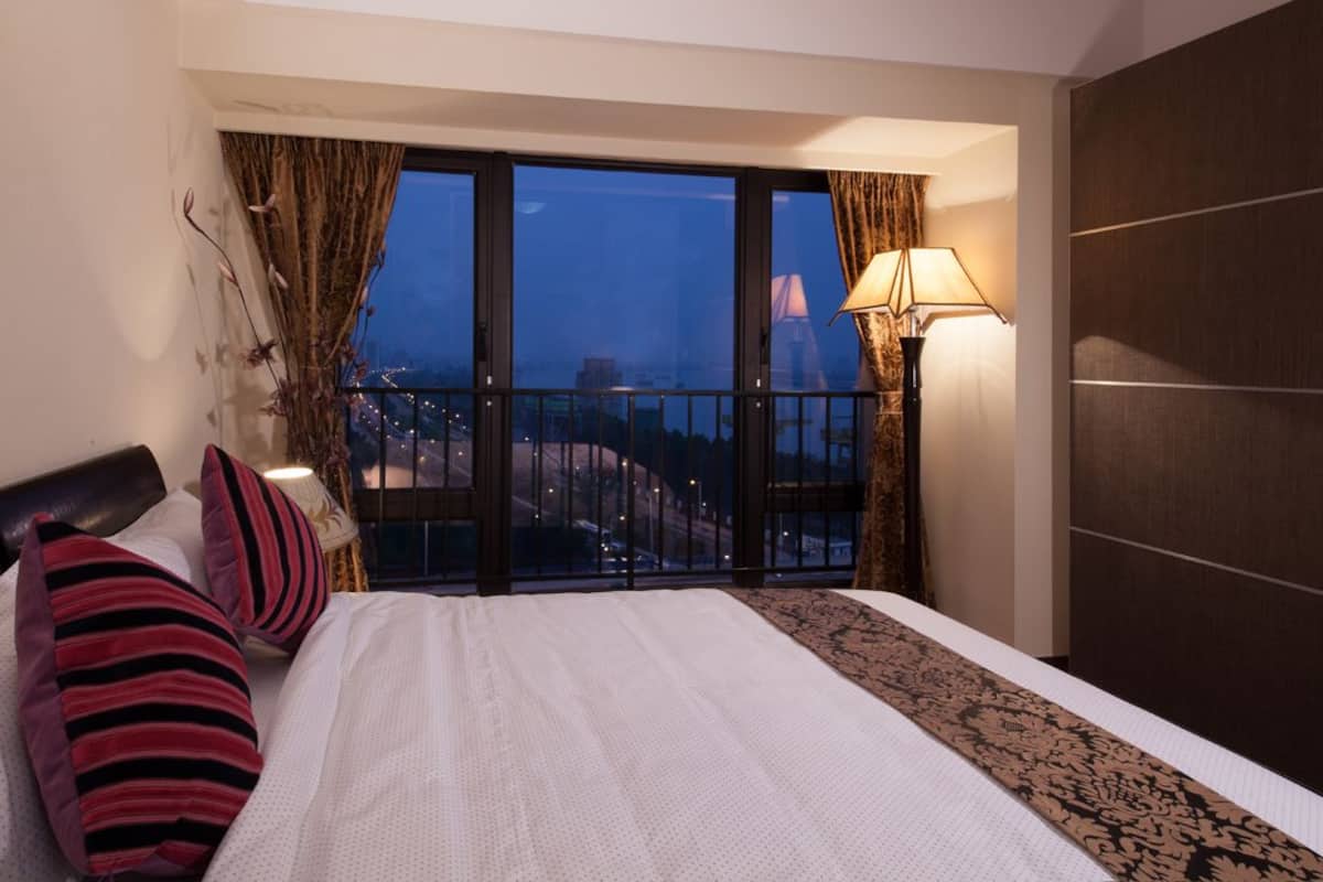 A hotel room with a large bed, a view of the cityscape through floor-to-ceiling windows, a nightstand with a lamp, and a dark-colored wardrobe.