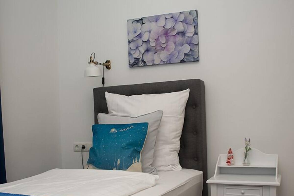 Ein ordentlich gemachtes Bett mit blauer Bettdecke, einem grauen Kopfteil und weißer Bettwäsche. Ein kleiner Nachttisch mit Lampe und einer Dekoration ist zu sehen.