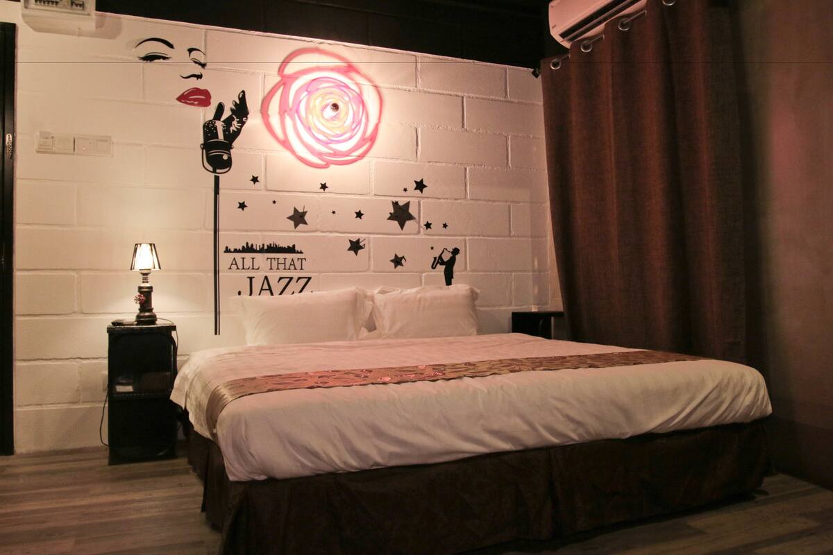 Un dormitorio con pared de ladrillo blanco, cama con ropa de cama blanca, mesita de noche con lámpara y decoración mural con estrellas y texto.