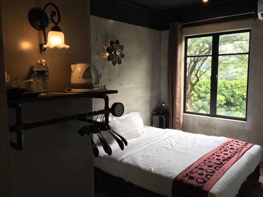 A bedroom with a bed, a shelf with a kettle, a water bottle, and a wall-mounted light fixture.