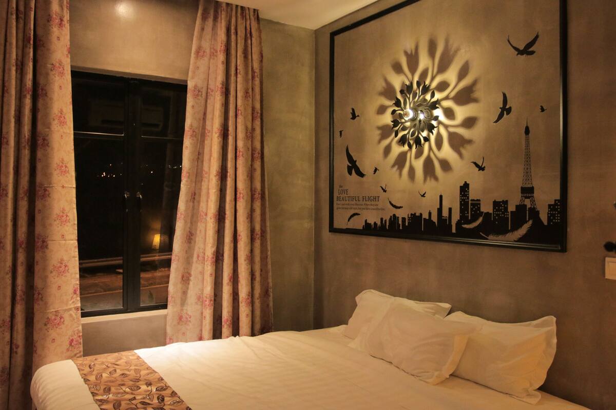 Habitación de hotel con cama, cortinas florales, una obra de arte con un paisaje urbano y ventana con vistas al paisaje urbano nocturno.
