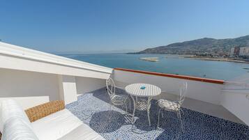 Un balcón con un sofá blanco, dos sillas y una pequeña mesa redonda. El balcón tiene un suelo de mosaico azul y da vista a una extensión de agua con una costa lejana.