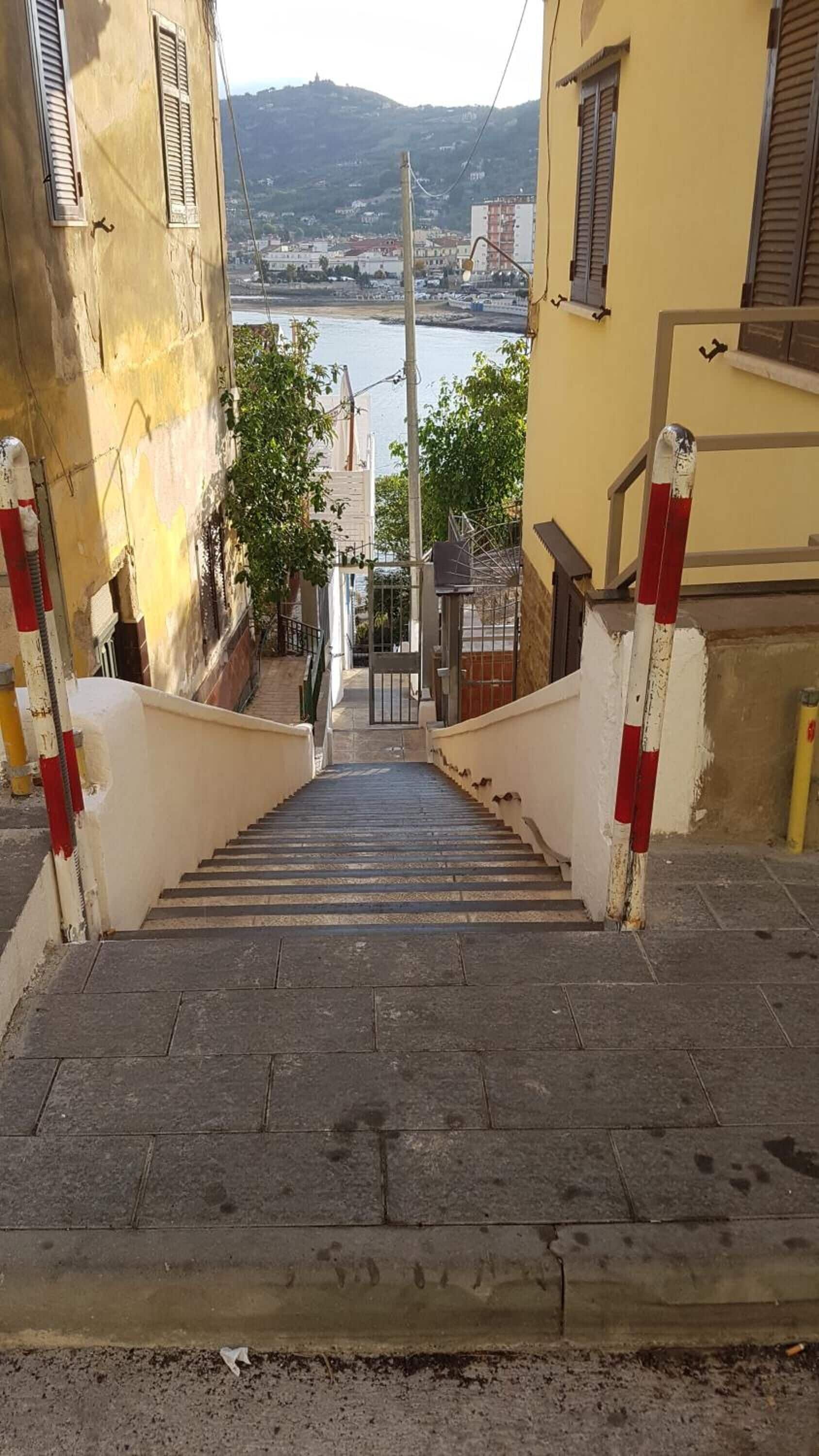 Una escalera conduce a una puerta, con edificios a ambos lados y una vista de una masa de agua y colinas al fondo.