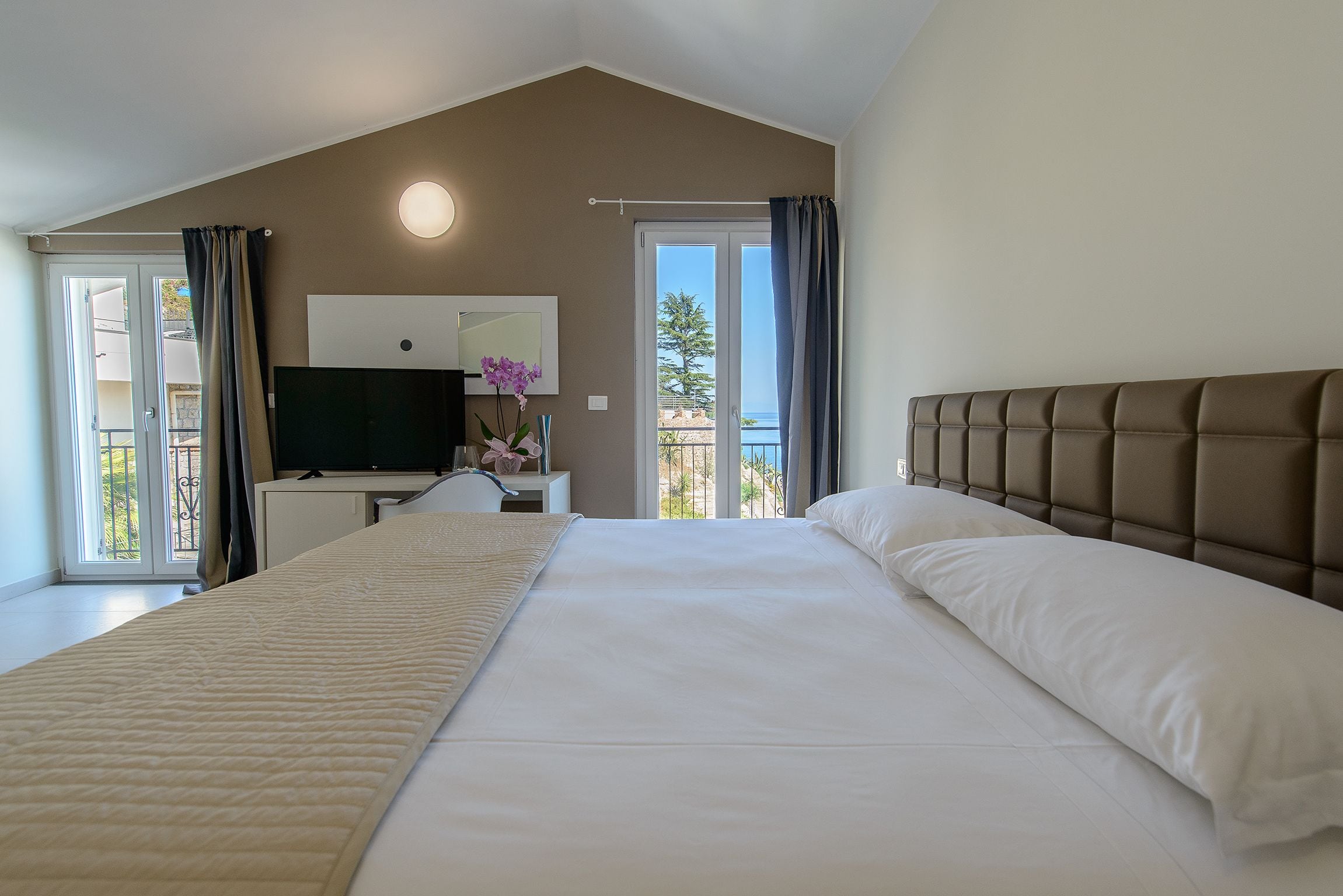 A hotel room with a large bed, a desk with a television, and a balcony with curtains.