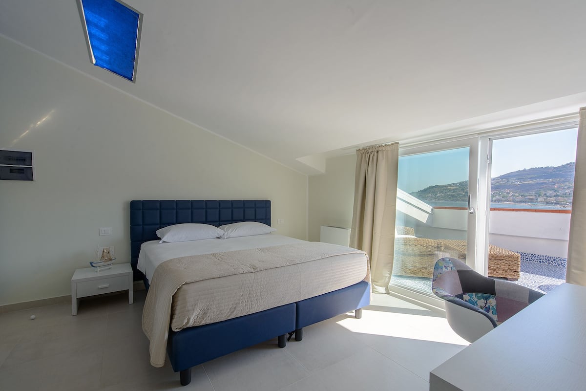 Ein Schlafzimmer mit einem großen Bett, einem Nachttisch, einem Sessel und Blick ins Freie.