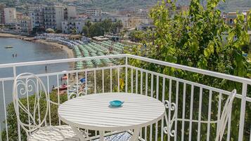 A balcony with a white metal railing, a round table, and two chairs. The balcony overlooks a beach with umbrellas and a view of the city and hills in the distance.